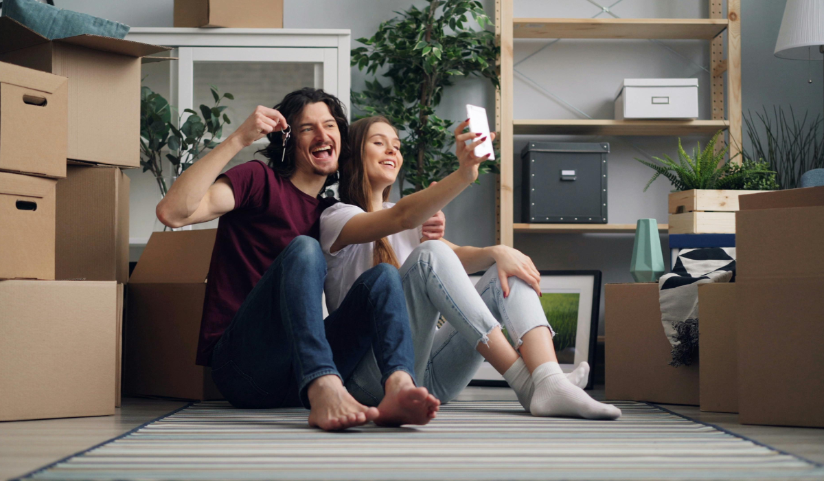 Un couple prend un selfie avec les clés de son nouveau logement, entouré de cartons de déménagement